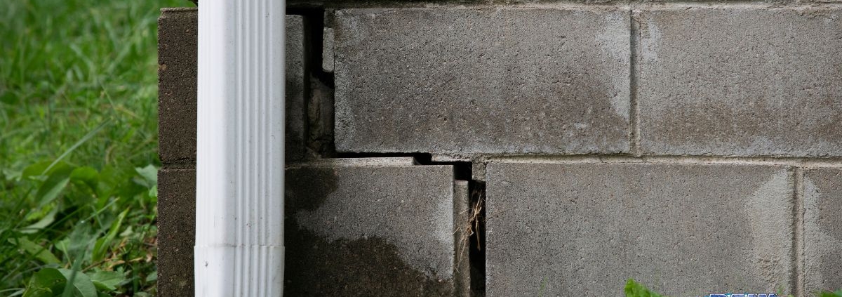close up of a brick foundation with a crack and a white gutter spout - Rain Master Seamless Gutters