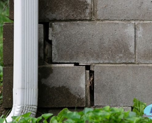 close up of a brick foundation with a crack and a white gutter spout - Rain Master Seamless Gutters